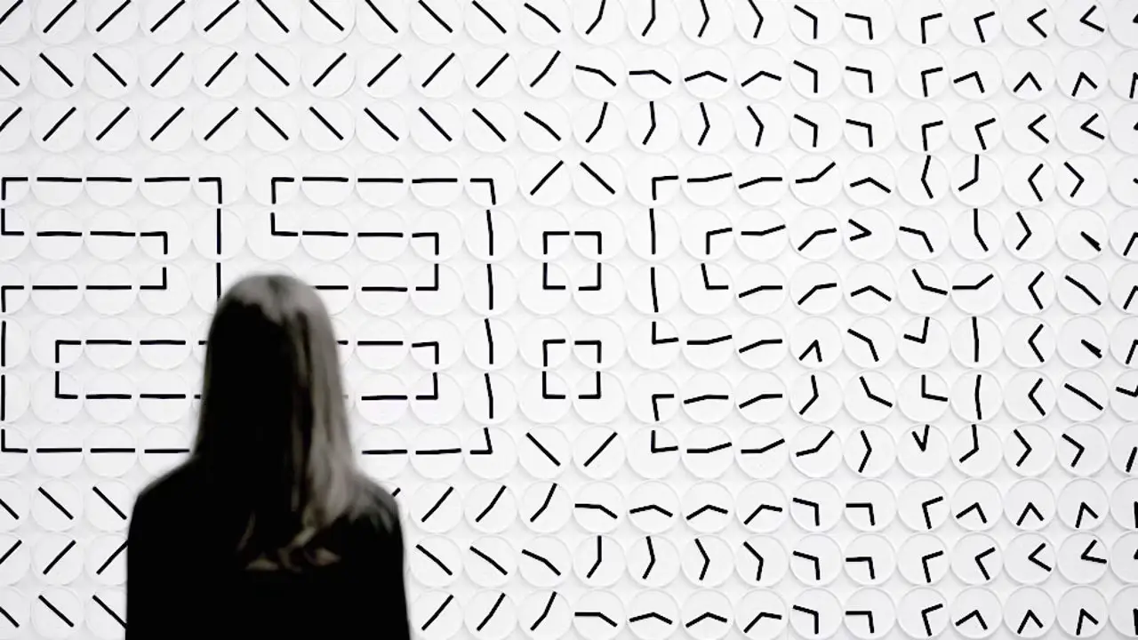 A visitor stands in front of A Million Times by Humans since 1982, a white wall of clocks whose black hands draw outlined digits, while the clocks around them are mid-turn