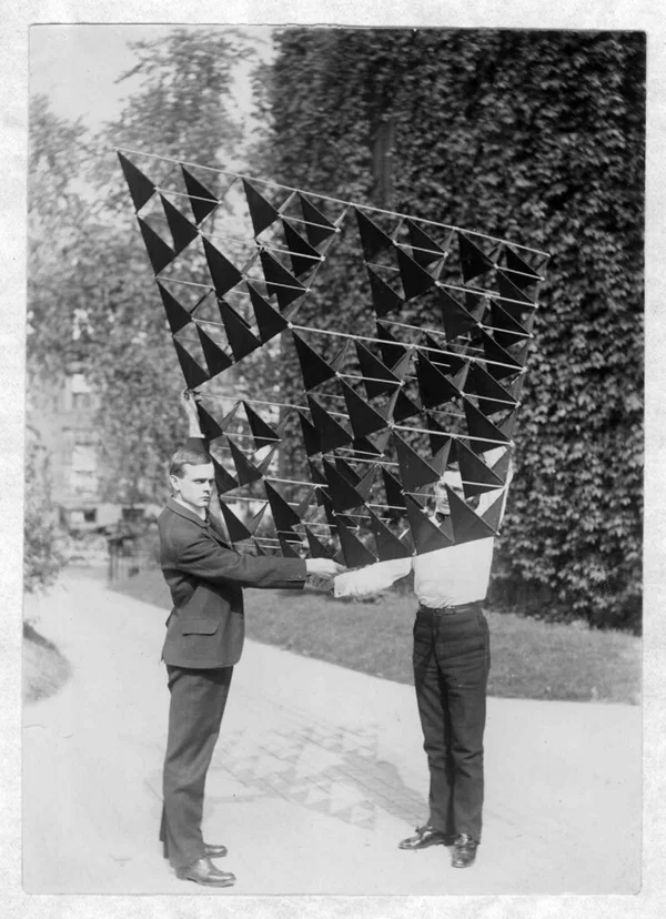 Two men carrying a silk-covered tetrahedral kite made of dozens of cells across a garden path