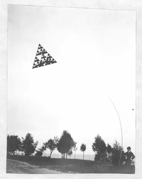 A 64-cell tetrahedral kite in flight, a dark Sierpiński triangle against a blank sky, with a man holding the line below
