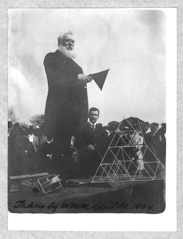 Alexander Graham Bell standing on a platform with a megaphone beside a bare tetrahedral kite frame, in front of a crowd, 30 April 1904