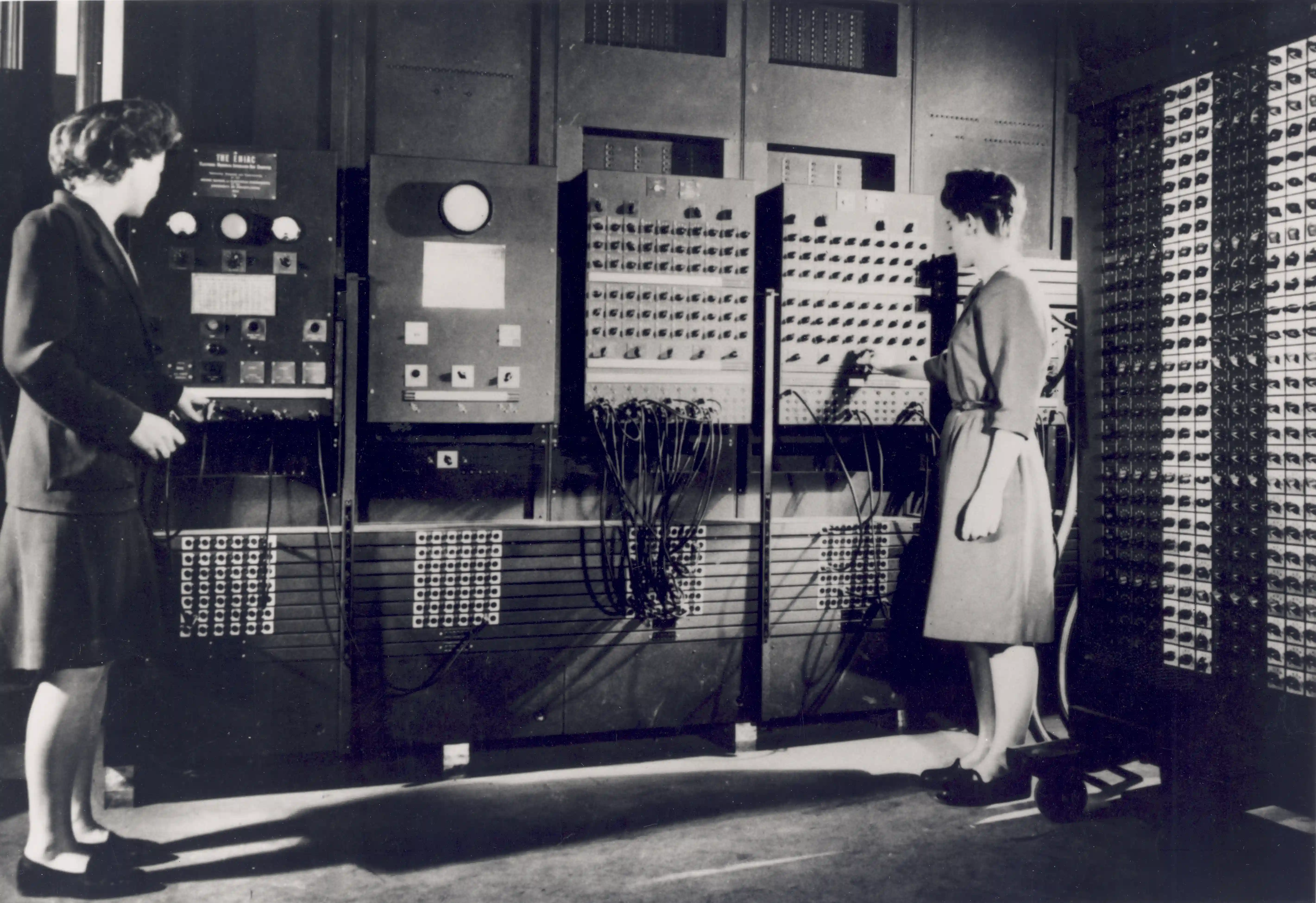 Black-and-white photograph of two women at ENIAC's control panels: walls of switches, dials and plugboards with bundles of cables, one woman setting a switch, the other watching a panel