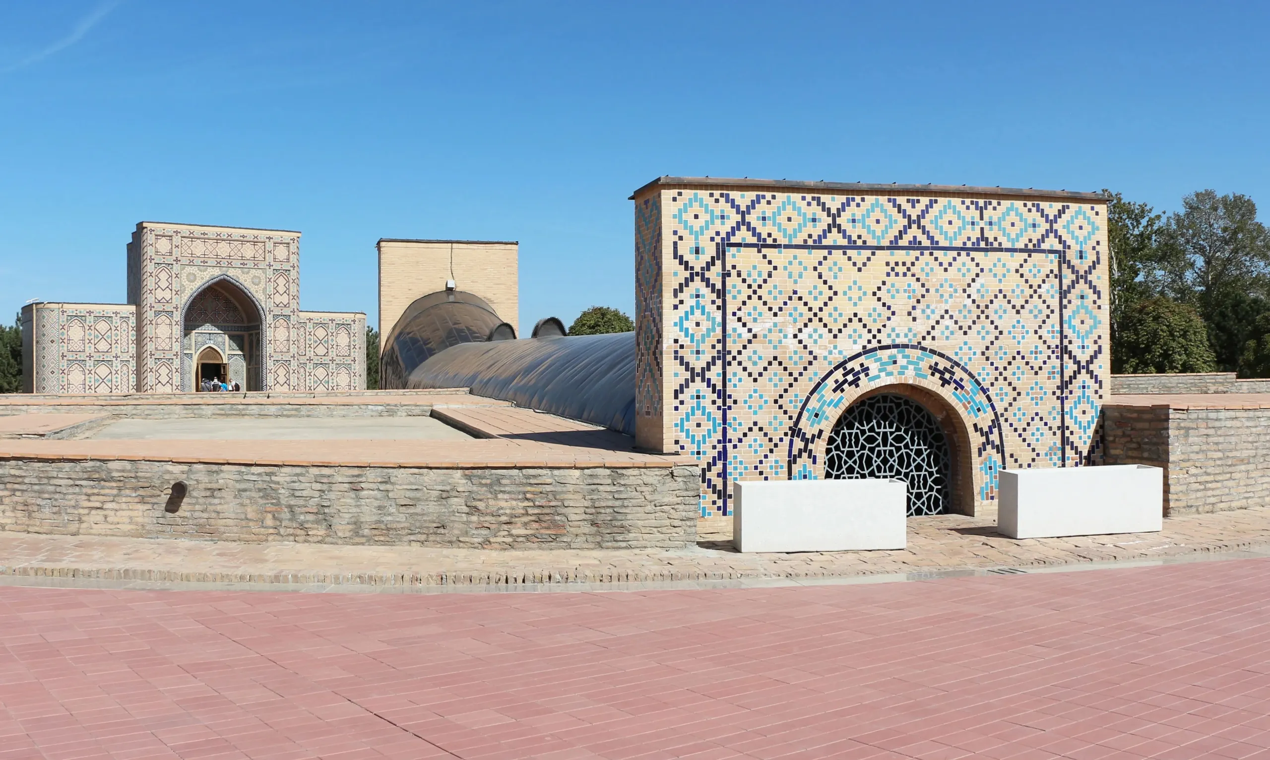 The Ulugh Beg Observatory site in Samarkand today: a tiled modern entrance with a lattice-covered arch, a long curved roof over a trench in the ground, and a restored portal behind it under a blue sky